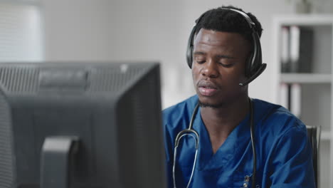 Afro-american-male-doctor-or-nurse-with-headset-and-computer-working-at-hospital-.young-professional-therapist-doctor-consulting-customer-client-using-remote-communication-speaking-on-webcam